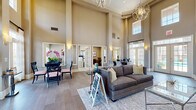 This single-floor community clubhouse or leasing center is defined by its dramatic double-height ceilings, which create an airy, grand ambiance throughout the main gathering area. Warm taupe and gray tones coat the walls, complemented by wide-plank wood-style flooring that runs seamlessly across the space. The main lounge area features a plush gray sofa, area rug, and accent tables beneath a striking geometric orb chandelier, establishing an inviting focal point for residents and visitors alike.

Adjacent to the lounge, a dining area hosts a sleek glass-top table with black chairs and vibrant orchid arrangements, adding a pop of color against the neutral palette. Tall French doors and transom windows line the perimeter, flooding the interior with abundant natural light while offering views to the outdoors.

A cozy sitting nook is anchored by a fireplace with rich wood built-ins displaying decorative accents, a wall-mounted television, and a patterned accent chair. Nearby, a dedicated workspace features a wood writing desk with chair, ideal for focused tasks. A stylish beverage station with a coffee maker provides a welcoming amenity. Private office spaces enclosed by glass-paned French doors are visible along the perimeter, supporting administrative or leasing functions. The overall atmosphere is polished, functional, and warmly welcoming.