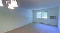 This single-floor space presents a clean, minimalist interior characterized by white walls and wall-to-wall carpeting in a neutral tone. The room is well-lit, benefiting from natural light that streams through a sliding double-pane window, which frames a view of lush green foliage outside. The window provides both visual connection to the outdoors and a sense of openness within the space. A baseboard heater runs along the exterior wall beneath the window, offering a practical heating solution. Overhead, a simple pendant light fixture provides additional illumination, complementing the natural light. The smooth, white ceiling and crisp wall surfaces contribute to a bright, airy ambiance throughout. The open layout offers flexibility for a variety of uses, with no obstructions limiting the arrangement of furnishings. The overall aesthetic is understated and neutral, providing a versatile canvas suitable for a range of interior design preferences. The space feels calm and functional, with practical features integrated seamlessly into its straightforward design.