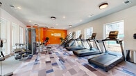 The Gibson Fitness Center at 2422 S Carrier Pkwy in Grand Prairie, Texas is a thoughtfully designed, single-floor facility that provides residents with a comprehensive workout environment. The space is divided into two distinct areas, each serving a specific fitness purpose.

The main cardio and strength zone is immediately recognizable by its bold orange accent wall, prominently branded with "The Gibson" logo, which adds a lively, energetic character to the room. This area houses a row of treadmills, stair climbers, elliptical machines, and a stationary spin bike, all arranged to maximize open floor space. Large windows along the exterior walls invite abundant natural light and offer views of the surrounding community.

The secondary area is dedicated to free weights and functional training. It features multiple dumbbell racks stocked with a full range of weights, adjustable workout benches, medicine balls, and a multi-functional cable machine suitable for a wide variety of strength exercises. Full-length wall mirrors expand the visual depth of the room and allow users to monitor their form during workouts.

Throughout both areas, a geometric, multi-toned carpet in shades of blue, grey, and white covers the floors, contributing to the modern aesthetic of the space. Recessed and flush-mount ceiling lighting ensures consistent illumination across the entire facility. The overall ambiance is bright, clean, and motivating, reflecting a well-maintained fitness environment.