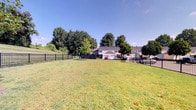 Located at 7999 Wildflower Ln in Westerville, Ohio, this single-level outdoor community space offers a generous open-air green area ideal for recreation and relaxation. The space is characterized by a well-maintained, expansive grassy lawn that provides ample room for a variety of outdoor activities. A sturdy black iron perimeter fence encloses the area, offering both definition and a sense of security while maintaining clear sightlines to the surrounding environment. Mature, leafy trees line the edges of the space, providing natural shade and a pleasant, park-like atmosphere. A gated entry point offers convenient access, and a seating or grilling area is visible near the fence line, making the space well-suited for casual gatherings. The backdrop features residential homes with attached garages, integrating this green space naturally within the neighborhood setting. Under open skies and surrounded by lush greenery, the space enjoys abundant natural light throughout the day, contributing to a welcoming and vibrant outdoor environment that serves as a communal hub for the surrounding residential community.