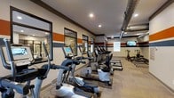 This fitness center is designed to deliver a comprehensive workout experience within a thoughtfully appointed space. The room features a striking contemporary aesthetic, highlighted by bold horizontal color-block striping in orange, dark brown, and beige along the upper walls, lending the space a dynamic, energetic character. Large windows along one side flood the area with natural light, while recessed ceiling lighting ensures consistent illumination throughout.

A lineup of modern cardio equipment — including elliptical trainers, upright and recumbent cycles, and treadmills — is arranged in an orderly row, each machine equipped with individual digital display consoles for tracking workout metrics. Exposed ductwork runs along the ceiling on one side, contributing an industrial accent that complements the space's modern design. A wall-mounted flat-screen television is positioned toward the far end of the room for entertainment during workouts.

The far end of the space transitions into a dedicated strength training area, where cycling bikes and additional equipment are visible. Stone-accented wall panels and large mirrors enhance the room's visual depth, making the space feel open and expansive. The flooring consists of carpet tiles that provide comfort underfoot, practical for the variety of equipment and activities the center accommodates.