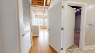 This two-floor space masterfully combines historic industrial character with contemporary updates, creating a distinctive and versatile environment. The upper level features warm hardwood flooring, exposed brick walls, and raw timber beam ceilings with visible ductwork, lending an authentic loft aesthetic. Large windows fitted with blinds allow generous natural light to fill the space, highlighting the textured materiality throughout. The kitchen on this level is equipped with wood cabinetry, a dark countertop, and standard appliances, set against an exposed brick backdrop framed by expansive windows. A hallway connects multiple rooms, with white doors providing clean contrast to the rustic surroundings. An in-unit stacked washer and dryer is tucked conveniently beside an original brick wall. The bathroom offers a tub-shower combination, wood vanity with dark countertop, and tile flooring. The lower level continues the industrial aesthetic with polished concrete floors, a striking exposed brick column, and a soaring wooden plank ceiling accented by a ceiling fan. This floor also includes a spacious walk-in closet with wire shelving and concrete flooring, providing ample storage.