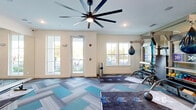 Located in Palm Coast, Florida, this fitness facility offers a thoughtfully designed exercise room that blends functionality with modern aesthetics. The space is finished with eye-catching herringbone-patterned carpet tiles in a palette of teal, gray, and blue, providing both visual appeal and a comfortable workout surface throughout the open floor area.

At the heart of the room are two RAX multi-functional training systems, each equipped with punching bags, suspension straps, resistance accessories, and built-in digital display screens, enabling a wide variety of strength and conditioning workouts. Dedicated workout mats are positioned in front of each station, creating organized training zones. Cardio equipment, including rowing machines, complements the functional training setup.

A large-scale ceiling fan anchors the room overhead, ensuring consistent air circulation during workouts. Multiple windows along the exterior walls invite ample natural light and offer views of the surrounding outdoor greenery, fostering an energizing atmosphere. A full-length wall mirror expands the sense of space and supports form-checking during exercise.

An accent wall featuring a soft botanical mural adds a distinctive design touch, while frosted glass interior doors provide access to adjoining spaces. The clean white walls and recessed lighting contribute to a bright, welcoming environment well-suited for a focused and effective fitness experience.