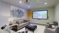 Located at 31611 Plaza De Toros Dr. in Spanish Fort, Alabama, this single-story home offers a thoughtfully designed living environment with standout entertainment amenities. The highlight of the space is a dedicated media room outfitted with a ceiling-mounted projector and a large-format projection screen, creating a true home theater experience. The room is finished with recessed lighting that allows for full ambiance control, neutral-toned walls, and crown molding that adds a refined architectural detail. Comfortable seating, including a plush sofa and upholstered ottoman, invites relaxed viewing. A large piece of abstract wall art in blue and white tones adds a bold decorative focal point, complementing the room's contemporary aesthetic. The light-colored flooring and soft area rug contribute to the room's warm, inviting atmosphere. The overall design of the space balances functionality with style, making it well-suited for both casual gatherings and dedicated entertainment. The single-floor layout offers convenient, accessible living throughout, with the media room serving as a central feature that elevates the home's overall appeal.