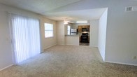 This single-floor residence offers a practical and functional layout centered around an open-concept living and kitchen area. The spacious living room is finished with wall-to-wall carpet flooring in a neutral tone, creating a warm and comfortable atmosphere. A ceiling fan with an integrated light fixture is centrally positioned overhead, providing both air circulation and ambient lighting. Natural light filters into the room through multiple windows, one of which is dressed with vertical blinds, adding to the bright and airy feel of the space. The textured ceiling adds a classic residential touch throughout. The kitchen is seamlessly connected to the living area and is separated by a raised breakfast bar counter, creating a natural transition between the two spaces. It is equipped with warm-toned wooden cabinetry, a black gas range, a black microwave mounted above, and a stainless steel refrigerator. A fluorescent light fixture illuminates the kitchen workspace, ensuring ample task lighting. The tile flooring in the kitchen contrasts neatly with the carpeted living area, clearly defining each functional zone. Overall, the space presents a clean, straightforward, and livable layout well-suited for everyday comfort.