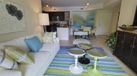 This single-floor space showcases a thoughtfully designed living area that combines coastal elegance with comfortable, inviting design. The room is anchored by a large sliding glass door with an arched transom window above, flooding the interior with natural light and framing picturesque waterfront views of a calm lake or canal beyond. Sheer white curtains frame the door, softening the bright outdoor light while maintaining an open, airy feel.

The living area is furnished with plush white sofas and a textured accent chair, all accented with coordinating throw pillows in soft shades of aqua, sage green, and cream — a palette that reinforces the coastal theme throughout. A vibrant striped area rug in greens and blues grounds the seating arrangement, adding warmth and visual interest underfoot.

Two round accent tables with distinct bases — one dark wood, one sculptural white — serve as stylish focal points within the arrangement. A tall floor lamp provides warm ambient lighting, complementing the soft wall art displayed nearby. The tile flooring, visible near the entryway to the outdoor space, adds a polished, practical element suited to the waterfront setting. Overall, the space exudes a relaxed yet refined coastal ambiance.
