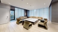 Located at 65 Broadway Ave in midtown Toronto, this contemporary amenity space is thoughtfully designed to offer residents a refined setting for both relaxation and social gatherings. The single-floor layout is divided into two distinct yet harmoniously connected zones.

The lounge area is elegantly appointed with plush, earth-toned sectional sofas and sculptural accent chairs arranged around a large marble-top coffee table. Floor-to-ceiling sheer drapery lines the expansive window wall, flooding the space with soft, diffused natural light and creating an airy, welcoming atmosphere. A wall-mounted flat-screen television and textured grey wall coverings add a polished, residential feel, while light wood-toned flooring ties the entire space together.

Transitioning through a corridor, the dining and kitchenette area is equally impressive. A long communal dining table surrounded by contemporary upholstered chairs serves as the focal point, anchored by a striking sculptural pendant light fixture. The kitchenette runs along one wall, featuring a sleek white countertop, integrated cabinetry, a sink, and a beverage refrigerator. A dramatic textured wood mosaic feature wall adds warmth and visual depth, creating a striking contrast against the clean, modern finishes throughout.