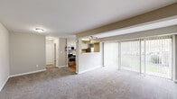 This single-floor unit offers a practical and well-organized layout comprising a combined kitchen and living room, one bedroom, a closet, a full bathroom, and a connecting hallway. The open-concept living area is carpeted and receives natural light through a large sliding glass door fitted with vertical blinds, providing outdoor views and direct access to the exterior. The entry door opens into the spacious living area, which flows visually into the semi-open kitchen separated by a half-wall partition with overhead track lighting.

The kitchen features white cabinetry, light speckled countertops with a single-basin stainless steel sink, hardwood flooring, a stainless steel refrigerator, and a freestanding stainless steel gas range. A window above the counter area allows additional natural light into the kitchen. The bedroom is a generously proportioned room with carpet, a ceiling fan with integrated lighting, and its own sliding glass door with vertical blinds offering outdoor views and natural light. The bathroom includes a full-sized tub and shower combination with white surround tiling, clean white fixtures, and a toilet. The neutral warm-beige wall tones and white trim throughout give the unit a cohesive, understated aesthetic.