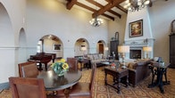 The Great Room presents a grand, open gathering space defined by its striking Spanish Colonial-inspired architecture. Soaring ceilings showcase exposed dark wood beams that run the length of the room, complemented by elegant wrought-iron and glass chandeliers that cast warm, ambient light throughout. A series of arched doorways line the perimeter, adding architectural rhythm while connecting the space to adjoining areas and allowing natural light to filter in from multiple directions. Upper clerestory windows further enhance the brightness of the room.

The focal point of the room is a stately white fireplace framed by tall, dark wood cabinetry and decorative artwork, flanked by ornate floor lamps and candlestick accents. Multiple seating arrangements, featuring rich leather and patterned upholstered sofas paired with dark wood coffee and side tables, are thoughtfully positioned to encourage conversation and relaxation. A grand piano is situated in one corner, adding a refined, cultural element to the space.

The boldly patterned, medallion-motif flooring in warm reds and golds anchors the entire room and reinforces the Mediterranean aesthetic. A round dining table with high-back chairs and fresh floral arrangements adds a welcoming, social dimension. The overall ambiance is warm, elegant, and inviting.