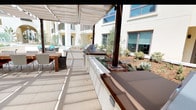 This well-appointed outdoor courtyard is designed for communal gathering and al fresco entertaining within a modern residential complex. The space is anchored by a generously sized covered pergola structure fitted with retractable canvas shade canopies supported by dark bronze-finished steel posts, providing adjustable sun protection throughout the day. Beneath the pergola, multiple built-in outdoor kitchens are outfitted with stainless steel gas grills, prep counters finished in rich dark wood-tone surfaces, and ample cabinetry — offering everything needed for outdoor cooking. Large wooden dining tables paired with houndstooth-patterned chairs and wicker seating accommodate multiple groups simultaneously. Beyond the covered area, an open-air lounge zone features sculptural wicker lounge chairs and a sectional sofa with colorful accent cushions arranged around low coffee tables, ideal for relaxed socializing. Raised planter beds filled with drought-tolerant landscaping including agave, ornamental grasses, and olive trees line the perimeter, adding a naturalistic, textured aesthetic. The light-colored large-format tile flooring throughout reflects natural sunlight, contributing to the bright, airy atmosphere of the courtyard.