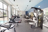 Located at 7931 Patriot Drive in Annandale, VA, this fitness center offers a thoughtfully designed exercise space built to support a wide range of workout routines. The single-floor layout is open and efficiently organized, providing ample room to move between stations without feeling cramped.

The exercise room is lined with large, floor-to-ceiling windows along multiple walls, flooding the space with natural light and offering views of the surrounding outdoor areas, including a terrace and neighboring greenery. The bright, airy atmosphere is complemented by a clean white ceiling fitted with track-mounted spotlights, while bold blue accent walls add a modern, energetic touch to the interior design.

Durable speckled rubber flooring runs throughout, providing cushioning and traction ideal for high-activity use. The cardio section features a row of treadmills, elliptical trainers, stationary bikes, and cycling machines equipped with individual display screens. The strength training area includes cable machines, a functional trainer, and multi-station weight equipment, all positioned along mirrored walls that enhance the sense of space. A dedicated stretching and open-floor zone rounds out the facility.

A wall-mounted climate control unit ensures comfort year-round. The overall design balances functionality with a clean, contemporary aesthetic, making this fitness center a well-appointed and practical space for daily exercise.