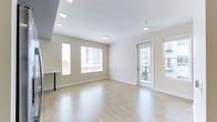 This contemporary single-floor space offers a clean, modern aesthetic defined by an abundance of natural light and thoughtful design details. The main living area is finished with smooth light hardwood flooring that reflects the natural light pouring in through multiple large windows fitted with blinds, creating a bright and airy atmosphere throughout. Recessed lighting in the ceiling supplements the natural light, ensuring the space is well-illuminated at any time of day. A ceiling fan adds both functionality and a subtle design element to the living area. A private balcony is accessible through a glass-paneled door, offering views of the surrounding urban environment and extending the sense of openness beyond the interior. The walls are finished in a soft, neutral tone that complements the light flooring and contributes to the overall clean and inviting ambiance. A built-in counter or bar-height surface near the window area provides a practical workspace or casual dining option. The layout is open and versatile, making efficient use of the available space while maintaining a comfortable, uncluttered feel. Overall, this unit presents a well-appointed, light-filled living environment with modern finishes and convenient urban-facing outdoor access.