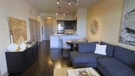 This single-floor residence showcases a thoughtfully designed open-concept layout that seamlessly connects the kitchen, dining, and living areas into one cohesive and inviting space. Rich, dark hardwood flooring runs throughout, anchoring the interior with warmth and sophistication.

The kitchen is a functional centerpiece, featuring dark espresso cabinetry, stainless steel appliances, and a granite-topped peninsula that doubles as a breakfast bar. Pendant lighting above the peninsula adds a refined touch, while track lighting on the ceiling ensures the entire space is well-lit.

Adjacent to the kitchen, the dining area accommodates an intimate round table with upholstered chairs, ideal for everyday meals or entertaining guests. A decorative sunburst wall mirror serves as a standout accent, lending an artistic focal point to the space.

The living area is generously proportioned, anchored by a large sectional sofa and complemented by a light-colored area rug that contrasts beautifully with the dark floors. The layout promotes easy flow between zones, making the space feel open and connected.

A hallway visible from the main living area leads to additional rooms, suggesting a well-organized floor plan designed for both comfort and practicality. The overall ambiance is modern, clean, and stylishly appointed.