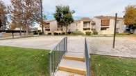 This residential complex is composed of several two-story buildings finished in a combination of stucco and horizontal lap siding in neutral tan and gray tones, accented by brown trim and low-pitched gabled rooflines. The exterior architecture is practical and consistent across the buildings, with balconies on upper-level units and large windows providing natural light throughout.

The grounds are thoughtfully maintained, featuring expansive open lawn areas interspersed with mature deciduous trees, tall palms, and neatly trimmed shrubs. Concrete walkways wind through the property, connecting the buildings and offering easy pedestrian access throughout the complex. Landscaped plant beds line the sides of buildings, adding greenery at ground level.

A paved court area — likely used for recreational activity — is situated centrally within the grounds, bordered by grass. Covered carport-style parking structures are visible toward the perimeter of the property, providing sheltered vehicle storage for residents. Accessible concrete steps with metal safety railings offer convenient connections between grade changes across the site.

The overall ambiance of the complex is open and spacious, with the mature tree canopy providing shade and seasonal color. The layout encourages outdoor activity and movement between buildings, creating a community-oriented environment with a calm, suburban character.