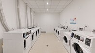 This shared laundry facility is a thoughtfully designed, single-floor space that offers a clean and functional environment for laundry needs. The room is finished with crisp white walls and a drop ceiling fitted with recessed lighting, creating a bright and well-lit atmosphere throughout.

Rows of commercial-grade, front-loading washing machines and dryers are arranged along both sides of the room, maximizing the use of floor space and providing ample capacity for multiple users. Each machine is clearly numbered for easy identification, and instructional signage is posted on the walls for user convenience. An electrical panel is also accessible within the room.

The flooring consists of durable, light-toned speckled linoleum, practical for a high-traffic laundry environment. A central floor drain is positioned in the middle of the room, reflecting a functional and safety-conscious design. Ventilation ducts run along one wall, ensuring proper exhaust for the dryer units. A baseboard heater provides climate control, keeping the space comfortable year-round.

A single entry door with a wood-paneled inset provides straightforward access to the room. Overall, the space is well-organized, meticulously maintained, and designed to accommodate shared use efficiently.