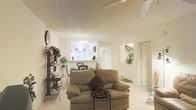 This single-floor space presents an open, flowing layout that seamlessly connects the living and dining areas. The interior is finished in crisp white tones across the walls and ceiling, contributing to a light, airy atmosphere throughout. Light-toned tile flooring runs continuously through the main living area, reinforcing the sense of openness and easy flow between spaces.

The living area is anchored by a cushioned sofa paired with a small accent table, providing a comfortable and functional seating arrangement. Decorative shelving along one wall adds character, displaying greenery and accessories that introduce warmth to the neutral surroundings.

Beyond the living area, the dining space is defined by a chandelier-style light fixture and benefits from natural light through a window that offers a view of outdoor greenery. This brightens the dining nook considerably and creates a welcoming atmosphere for meals.

A hallway leads to additional rooms, with doorways visible from the main living space. Wall niches with decorative plant arrangements punctuate the transition areas, adding subtle visual interest. Overall, the space maintains a clean, well-lit, and cohesive aesthetic across its single level.