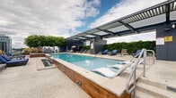 Located at 1575 Anderson Rd in McLean, Virginia, this rooftop pool deck offers a sophisticated outdoor retreat with sweeping views of the surrounding McLean and greater Washington, D.C. metro skyline. The centerpiece of the space is a generously sized, crystal-clear swimming pool with a sleek, modern design, complemented by an adjacent raised spa featuring a cascading water feature with warm wood-toned cladding and stone tile surround. A contemporary louvered pergola structure spans a significant portion of the deck, providing adjustable shade while maintaining an open, airy atmosphere. The pool deck is well-appointed with ample blue chaise lounges and lounge chairs, as well as additional seating areas with woven club chairs, offering a variety of relaxation zones. A covered pavilion area houses a bar-height dining setup enclosed by glass railings, ideal for social gatherings. The perimeter of the deck is lined with mature hedging and ornamental trees, lending a sense of privacy and greenery against the urban backdrop. Crisp, light-toned concrete decking ties the space together with a clean, modern aesthetic. The overall ambiance is that of a refined, resort-inspired rooftop amenity designed for both relaxation and socializing.