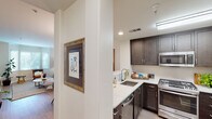 This single-floor residence offers a thoughtfully designed layout that balances functionality and style throughout its interconnected living spaces. The kitchen is a standout feature, showcasing rich dark-stained cabinetry with sleek hardware, light quartz countertops, and a full suite of stainless steel appliances including a gas range, over-the-range microwave, and dishwasher. A half-wall pass-through connects the kitchen seamlessly to the dining area, which is anchored by a warm wood round table beneath an elegant multi-pendant chandelier. Light hardwood-style flooring flows through the main living areas, enhancing the open, airy feel of the space. The living room is a welcoming retreat, highlighted by a graceful bay window fitted with plantation shutters that invite abundant natural light. Crown molding frames the room, while built-in wall niches provide a unique architectural touch for display. The bedroom offers a serene atmosphere with soft carpeting and generous wall space. The bathroom features a wide vanity with a dark countertop, an undermount sink, and a large mirror, complemented by a window that provides natural light and a glimpse of surrounding greenery. Throughout the space, neutral warm tones create a cohesive and inviting ambiance.