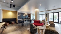 This 2-bedroom, 2-bathroom residence offers a thoughtfully designed single-floor layout that blends modern aesthetics with functional living. The open-concept common area serves as the heart of the space, seamlessly connecting the kitchen and living zones in a fluid, inviting arrangement. The kitchen features dark tile backsplash detailing, sleek cabinetry, and a stylish bar-height counter with contemporary seating, making it ideal for both casual dining and entertaining. Statement pendant lighting with a geometric, multi-arm design adds a dramatic focal point above the kitchen area, complementing the warm wood-toned flooring that runs throughout. The living area is furnished with an eclectic yet curated mix of seating, including a bold red curved sofa, patterned accent chairs, and gold-finished side tables, creating a dynamic and sophisticated atmosphere. A structural column is integrated seamlessly into the open floor plan, adding an architectural element to the space. Large windows allow natural light to filter in, offering views of the surrounding urban environment and enhancing the bright, airy ambiance. Recessed lighting and additional decorative fixtures ensure well-layered illumination throughout, completing this polished and contemporary residence.