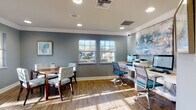 Located at 2042 Arrowgrass Dr in Wesley Chapel, Florida, this single-floor business center is thoughtfully designed to support both focused work and informal collaboration. The space is finished with warm hardwood-style flooring and cool blue-gray walls accented by crisp white crown molding, creating a professional yet welcoming atmosphere throughout.

The main work area features a dedicated computer station equipped with multiple desktop computers and a multifunction printer, set along a clean white desk against the wall. Ergonomic task chairs in teal and gray complement the modern aesthetic. A large, vibrant abstract painting anchors this workspace, adding a creative energy to the environment.

Adjacent to the workstation area, a dining-height table with patterned upholstered chairs provides a comfortable space for small group meetings or collaborative sessions. Multiple double-hung windows throughout the space invite generous natural light and offer views of the surrounding landscaped exterior.

A secondary seating nook features a bistro-style high table with bold blue bar stools alongside a leather bench, offering a casual spot for quick conversations or informal gatherings. Recessed lighting supplements the natural light, ensuring the space remains bright and functional at all times. The overall design balances productivity and comfort, making it a versatile and inviting environment.