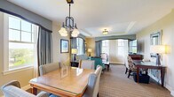 This elegantly designed one-bedroom suite offers a refined and comfortable living experience across a single, well-organized floor. The open layout connects the kitchen, dining area, and living room in a seamless flow, creating a welcoming and functional environment throughout.

The kitchen is outfitted with rich warm-toned cabinetry, granite countertops, a large island with a sink, stainless steel appliances including a full-size refrigerator, gas range, and built-in microwave, and tile flooring — providing everything needed for full meal preparation. Pendant lighting above the island adds a polished finishing touch.

The dining area accommodates a substantial wood dining table surrounded by upholstered chairs, illuminated by an elegant chandelier. Multiple large windows dressed with draped curtains allow abundant natural light and views of the surrounding landscape.

The living room features a comfortable sofa and accent chairs arranged around a coffee table, with additional windows framing scenic outdoor views, contributing to an airy and light-filled atmosphere.

The spacious primary bedroom is appointed with large windows on multiple walls, plush carpeting, and tasteful furnishings, creating a calm and restful retreat. The full bathroom includes a granite-topped vanity with warm wood cabinetry, tile flooring, a glass-enclosed shower, and a soaking tub.