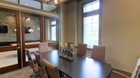 This single-floor conference room is designed to accommodate professional gatherings in a polished, well-appointed setting. The centerpiece is a large, dark-toned rectangular table surrounded by cushioned tan leather chairs with sleek metal frames, offering both comfort and a contemporary aesthetic. Tall windows along one wall allow abundant natural light to fill the space, providing views of surrounding greenery and contributing to an open, airy atmosphere. The elevated ceiling height enhances the sense of volume, and an industrial-style chandelier overhead adds a refined touch to the room's overall design. Neutral warm-toned walls complement the rich wood finishes and stone accent details visible through a glass partition, which separates this conference area from an adjoining space. The adjoining room, partially visible through the partition, features additional seating and architectural interest, including a stone accent wall and pendant lighting. The combination of natural materials, sophisticated furnishings, and generous natural light creates a professional yet inviting environment well-suited for meetings, presentations, or collaborative work sessions.