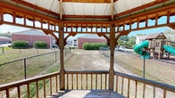 This outdoor amenity area in Hattiesburg, Mississippi centers around a well-constructed octagonal gazebo featuring warm stained wood framing, decorative bracketed trim, and a white metal roof that provides reliable shade and shelter. The gazebo is elevated on a wood deck platform enclosed by a sturdy wooden railing, with built-in bench seating along the interior perimeter—ideal for gathering or relaxing while taking in panoramic views of the surrounding grounds.

The open-sided design allows natural light and breezes to flow freely through the structure, while the decorative curved woodwork along the upper frame adds architectural character. A concrete walkway connects the gazebo to adjacent community facilities, including a fenced dog park visible to one side, enclosed with chain-link fencing and providing a dedicated space for pets. To another side, a playground is clearly visible, equipped with a multi-level structure featuring green tube slides and climbing elements set on a rubberized surface.

The broader grounds consist of open grassy areas, mature trees offering additional shade, and chain-link perimeter fencing. Together, these amenities form a practical and welcoming outdoor community space suited for residents of all ages.
