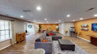 This single-floor community clubhouse is thoughtfully designed to offer a variety of functional and social spaces under one roof. The welcoming entrance features tile flooring that transitions into wood-look planks extending throughout the rest of the space, creating a cohesive and warm aesthetic. Recessed lighting and textured ceilings are consistent throughout, providing even illumination across each area.

The main lounge is generously proportioned, anchored by a large sectional sofa with tufted upholstery, colorful accent pillows, and a patterned area rug. Bold orange accent walls add energy and visual interest, a design choice carried consistently through multiple zones. An entertainment corner features a wall-mounted flat-screen television, floating shelves, and a classic popcorn machine, making it an ideal spot for casual gatherings.

A dedicated dining area accommodates a round table surrounded by ornate upholstered chairs, bathed in natural light from a window dressed with patterned curtains. Adjacent to the main lounge is a well-appointed kitchenette showcasing rich dark cabinetry, granite countertops, a stainless steel under-counter refrigerator, and a striking floor-to-ceiling linear mosaic tile backsplash. The space concludes with a business center alcove, rounding out this versatile and inviting communal facility.