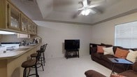 This single-floor residence offers an open and functional layout designed for comfortable everyday living. The living area serves as the heart of the space, featuring light-colored carpet flooring that adds warmth underfoot and a ceiling fan with an integrated light fixture that keeps the room well-ventilated and bright. A textured ceiling adds subtle architectural character to the room.

The living area flows naturally into a bar-style counter that separates it from the kitchen, creating a casual and inviting social zone. Bar stools line the counter, providing a convenient spot for casual dining or conversation. The kitchen cabinetry, visible beyond the counter, features light wood tones with glass-front upper cabinets, lending an open and airy feel to the space.

The living room is thoughtfully arranged with ample space for seating and entertainment, anchored by a wall-mounted television and accented by neutral walls that offer a versatile backdrop for a range of décor styles. The overall ambiance of the space is relaxed and welcoming, with a cohesive color palette of warm neutrals and deep browns that ties the rooms together seamlessly.