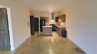 This single-floor space showcases an open-concept layout that seamlessly integrates the living and kitchen areas. The polished concrete floors extend throughout, lending an industrial yet modern aesthetic to the interior. The kitchen is a functional centerpiece, featuring warm-toned wood cabinetry that provides ample storage, a central island ideal for meal preparation or casual dining, and black appliances including a refrigerator and gas range. A prominent stainless steel wall-mounted range hood serves as a striking focal point above the cooktop, while a double sink with light-colored countertops lines the adjacent wall. Track lighting runs along the ceiling, offering directed illumination across the kitchen space. The open floor plan creates a generous, airy feel, with the living area flowing naturally from the kitchen. Black-painted doors add a bold contrast to the neutral cream-toned walls, contributing to the space's contemporary character. A visible HVAC vent near the entry door ensures climate control throughout. The overall ambiance is clean and industrial-modern, with a straightforward, practical design suited for everyday living.