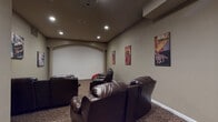 This single-floor theater room at 205 Atlantic Ave in Pittsburg, California, is designed to deliver an immersive cinematic experience. The focal point of the space is a large projection screen framed by an elegant arched architectural detail and a decorative column, lending a classic movie-house aesthetic to the room. Recessed lighting in the ceiling provides ambient illumination, while a ceiling-mounted projector ensures a high-quality viewing experience.

Seating consists of multiple dark brown leather reclining sofas and chairs arranged in a tiered layout facing the screen, with built-in cup holders for added convenience. The patterned carpet in warm earth tones contributes to the room's comfortable, lounge-like atmosphere. Movie-themed vintage-style art posters — referencing film, art, and cinema — are displayed along the taupe-toned walls, reinforcing the entertainment theme throughout.

A compact kitchenette is integrated into the back of the room, featuring wood-tone cabinetry, an under-counter refrigerator, and a sink, making it easy to prepare snacks and beverages without leaving the space. A doorway connects to an adjacent corridor, providing convenient access to additional areas. Overall, the room balances functional design with a stylish, theater-inspired ambiance.