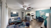 This single-floor residence offers a thoughtfully designed open-concept layout that seamlessly connects the living, dining, and kitchen areas. The living room features rich hardwood-style flooring, a ceiling fan, and a sliding glass door that opens to a private balcony, filling the space with natural light. A bold teal accent wall adds a vibrant coastal touch that is carried throughout the interior design.

The kitchen is well-equipped with white cabinetry, light countertops, a tile backsplash, black appliances including a refrigerator and range, and a breakfast bar with bar seating — ideal for casual dining. Adjacent to the kitchen, a dedicated dining area provides additional space for entertaining.

The primary bedroom is a comfortable retreat featuring plush carpet, a striking teal accent wall, ample natural light from multiple windows, and direct access to the outdoors. The en-suite bathroom includes a dual-sink vanity with white cabinetry and a separate walk-in shower. The second bedroom also features carpet flooring, bright windows with decorative curtains, and its own access to a full bathroom complete with a shower-tub combination. A linen closet and additional storage areas contribute to the overall functionality of this well-organized home.