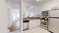 This single-floor residence offers a thoughtfully designed layout that balances functionality and comfort throughout. The kitchen is outfitted with white shaker-style cabinetry, dark granite countertops, light tile flooring, and a full suite of stainless steel appliances including a refrigerator, range, microwave, and dishwasher. A pass-through opening connects the kitchen to the main living area, enhancing the open feel of the space. Conveniently tucked adjacent to the kitchen is an in-unit laundry closet housing a full-size washer and dryer. The spacious living area features warm hardwood-style flooring, a ceiling fan with integrated lighting, and track lighting overhead. A sliding glass door leads directly to an enclosed screened lanai, which wraps around with floor-to-ceiling windows offering serene views of lush green grounds and surrounding landscaping. The bedroom is generously sized with plush carpet flooring and provides access to both a closet and the full bathroom. The bathroom includes a large vanity with dark granite countertops, dual mirrors, and bright overhead lighting, with tile flooring that extends through the hallway. Neutral tones and an abundance of natural light throughout lend the space a clean, airy ambiance.