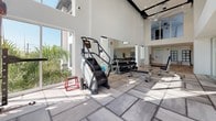This state-of-the-art fitness center is designed to accommodate a wide range of workout styles within a bright, contemporary setting. The facility is characterized by soaring double-height ceilings and floor-to-ceiling windows that flood the space with natural light while offering views of the surrounding greenery. Large ceiling fans are suspended overhead, enhancing airflow throughout the expansive workout areas.

The flooring transitions between light hardwood and large-format tile, subtly delineating distinct functional zones. A standout feature is the full-height rock climbing wall, accented with colorful handholds and set against a bold patterned backdrop, providing a unique vertical training option. Adjacent to it, a dedicated strength zone houses a Matrix functional training system, weight benches, and a comprehensive dumbbell rack displayed along mirrored walls.

The cardio zone is well-equipped with multiple treadmills, elliptical trainers, stair climbers, and Concept2 rowing machines arranged to maximize space and usability. A separate open-floor studio area with hardwood flooring offers ample room for stretching, yoga, or group fitness activities. The overall ambiance is clean, airy, and energizing, with a modern white palette and generous glazing creating an inviting environment for a comprehensive fitness experience.