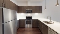 This single-story unit in Piedmont showcases a clean, contemporary design throughout. The open living area immediately draws attention with its impressively tall ceilings and crisp white walls, creating a bright and airy atmosphere. Dark-toned hardwood flooring extends seamlessly through the main living space, offering a striking contrast against the light surroundings. Large windows allow natural light to pour in, further enhancing the open feel of the space.

The kitchen is well-appointed with dark espresso-finished cabinetry extending to the upper walls, offering generous storage. A white subway tile backsplash pairs cleanly with light quartz countertops and a peninsula with an undermount sink. Stainless steel appliances, including a refrigerator, range, microwave, and dishwasher, complete this functional culinary space. Pendant lighting above the peninsula adds a refined touch.

The bedroom features plush carpeting, a natural light source through a tall window, and leads directly into a spacious walk-in closet outfitted with wire shelving and hanging rods. The full bathroom includes a soaking tub with shower, white tile surround, a dark wood vanity with a white countertop, and a large mirror.