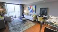This thoughtfully designed single-floor space makes a strong first impression with its modern aesthetic and carefully curated interior. The living area serves as the heart of the space, anchored by a crisp white sofa adorned with an eclectic mix of patterned and solid accent pillows in yellow, navy, and grey tones. A large, colorful abstract painting commands attention on the main wall, infusing the room with energy and artistic character. Warm hardwood flooring runs throughout, complemented by a textured area rug in cool grey and blue tones that defines the seating zone. A tufted ottoman doubles as a coffee table, adding both function and style. Natural light floods the space through floor-to-ceiling sliding glass doors that open onto a private balcony, offering views of lush, tree-lined surroundings. Patterned drapes in bold geometric prints frame the balcony entrance, adding visual depth. A sleek glass-top dining or work table with yellow upholstered chairs sits adjacent to the living area, creating a versatile zone for dining or productivity. A large decorative clock anchors the far wall, while recessed lighting ensures the space remains well-lit throughout. The overall ambiance is contemporary, vibrant, and inviting.