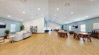 Located at 660 Aubrey Ln in Calabash, NC, this generously sized single-story facility offers an open, airy layout thoughtfully designed for communal gathering and relaxation. The space is defined by soaring vaulted ceilings in the main area, recessed lighting throughout, and warm light wood flooring that flows seamlessly from room to room.

The welcoming entrance foyer features glass double doors that flood the space with natural light, complemented by curated accent seating, a round wall mirror, and lush potted plants that set a coastal tone. The expansive main hall transitions effortlessly into distinct lounge and dining zones. The lounge area is furnished with plush neutral sofas, accent chairs, and a wall-mounted display, all anchored by soft blue accent walls that evoke a beachy, relaxed atmosphere.

The dining area accommodates multiple seating arrangements with mid-century modern wooden tables and chairs, and is supported by a kitchenette outfitted with white cabinetry, granite-style countertops, and a stainless steel refrigerator. A quieter sitting nook with cozy armchairs and coastal artwork provides a more intimate retreat. The space also includes two bathrooms and a closet, rounding out a well-appointed, versatile facility ideal for community events and social gatherings.