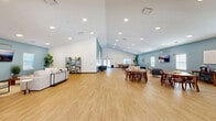 Located at 660 Aubrey Ln in Calabash, NC, this generously sized single-story facility offers an open, airy layout thoughtfully designed for communal gathering and relaxation. The space is defined by soaring vaulted ceilings in the main area, recessed lighting throughout, and warm light wood flooring that flows seamlessly from room to room.

The welcoming entrance foyer features glass double doors that flood the space with natural light, complemented by curated accent seating, a round wall mirror, and lush potted plants that set a coastal tone. The expansive main hall transitions effortlessly into distinct lounge and dining zones. The lounge area is furnished with plush neutral sofas, accent chairs, and a wall-mounted display, all anchored by soft blue accent walls that evoke a beachy, relaxed atmosphere.

The dining area accommodates multiple seating arrangements with mid-century modern wooden tables and chairs, and is supported by a kitchenette outfitted with white cabinetry, granite-style countertops, and a stainless steel refrigerator. A quieter sitting nook with cozy armchairs and coastal artwork provides a more intimate retreat. The space also includes two bathrooms and a closet, rounding out a well-appointed, versatile facility ideal for community events and social gatherings.