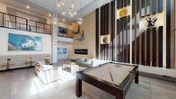 This expansive, open-plan space is defined by its dramatic double-height ceilings, which create an immediately striking sense of volume and grandeur. A cluster of globe pendant lights cascades from above, serving as a bold focal point while illuminating the main living area below. Large windows positioned at both ground and upper levels flood the interior with abundant natural light, enhancing the bright, airy atmosphere throughout.

The main floor is thoughtfully organized into several distinct zones. A central lounge area features a tufted velvet sofa paired with a contemporary coffee table, anchored by a large area rug. Adjacent to this is a full-size pool table, adding a recreational element to the space. A standout feature wall clad in herringbone-patterned wood paneling houses a sleek, wide linear fireplace with a floating hearth shelf, flanked by dark vertical slat detailing with decorative niches. Suspended sculptural figures add an artistic dimension to this wall.

Further into the space, a dining area with a round table and upholstered chairs sits alongside a lounge with leather seating and a wall-mounted television. A mezzanine level with cable railings overlooks the main floor, contributing to the space's layered, modern aesthetic. Large-format tile and hardwood flooring complete the polished interior design.