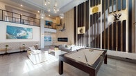 This expansive, open-plan space is defined by its dramatic double-height ceilings, which create an immediately striking sense of volume and grandeur. A cluster of globe pendant lights cascades from above, serving as a bold focal point while illuminating the main living area below. Large windows positioned at both ground and upper levels flood the interior with abundant natural light, enhancing the bright, airy atmosphere throughout.

The main floor is thoughtfully organized into several distinct zones. A central lounge area features a tufted velvet sofa paired with a contemporary coffee table, anchored by a large area rug. Adjacent to this is a full-size pool table, adding a recreational element to the space. A standout feature wall clad in herringbone-patterned wood paneling houses a sleek, wide linear fireplace with a floating hearth shelf, flanked by dark vertical slat detailing with decorative niches. Suspended sculptural figures add an artistic dimension to this wall.

Further into the space, a dining area with a round table and upholstered chairs sits alongside a lounge with leather seating and a wall-mounted television. A mezzanine level with cable railings overlooks the main floor, contributing to the space's layered, modern aesthetic. Large-format tile and hardwood flooring complete the polished interior design.