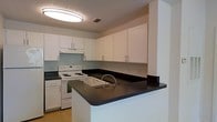 This single-floor residential space is designed with practicality and clean aesthetics in mind. The kitchen serves as a central highlight, showcasing a peninsula layout with dark granite-style countertops that contrast sharply against the bright white cabinetry. An abundance of upper and lower white shaker-style cabinets provides generous storage, keeping the space organized and visually uncluttered. The peninsula extends the counter space and doubles as a breakfast bar or prep area, making it a versatile feature for both cooking and casual dining. A stainless-steel arc faucet is positioned at the peninsula sink, adding a contemporary touch. The white electric range and matching appliances contribute to the cohesive, clean look of the kitchen. A flush-mounted ceiling light fixture illuminates the space with even, ambient lighting throughout. The light-toned tile flooring complements the overall neutral color palette, while the textured ceiling adds a subtle architectural detail. The open-concept design between the kitchen and adjacent living areas enhances the sense of flow and spaciousness throughout the single-level floor plan, making it a well-appointed and functional living environment.