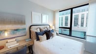 This bedroom is thoughtfully designed with a clean, contemporary aesthetic that balances comfort and style. The room is anchored by a generously sized bed with a woven, arched headboard featuring nailhead trim detailing, dressed in crisp white bedding and layered with an assortment of navy, charcoal, and neutral-toned accent pillows. A sleek wooden nightstand sits alongside the bed, providing functional surface space with drawer storage beneath.

The walls are finished in a soft, muted sage-white tone that contributes to the room's serene and airy feel. Framed artwork and prints displayed above the headboard add a subtle decorative touch without overwhelming the space. Light wood flooring complements the overall neutral palette and enhances the sense of warmth throughout.

A standout feature of the room is the large, floor-to-adjacent-ceiling window framed in black, which floods the space with natural light and offers views of the surrounding urban landscape. The window's modern black framing adds a sharp, architectural contrast to the room's otherwise soft palette, lending a contemporary edge to the overall design.