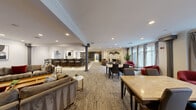 The Arrive Stamford Lounge is a thoughtfully designed, single-floor community space that blends functionality with refined style across several distinct zones. The main gathering area is anchored by a white-brick fireplace framed by decorative wall art and crown molding, surrounded by plush seating including rich red accent chairs and neutral-toned sofas arranged on textured carpeting. Large multi-pane windows along one wall flood the space with natural light and offer views of the outdoor surroundings, framed by floor-length linen drapes. Architectural columns punctuate the open layout, adding a classic structural character throughout.

A full-service kitchen occupies one end of the lounge, outfitted with gray cabinetry, dark granite countertops, stainless steel appliances including a range with a statement tile backsplash and overhead hood, and a breakfast bar with seating. Adjacent to the kitchen, a darker-toned bar area with counter-height stools provides an additional social hub.

A dedicated game room features light hardwood flooring, a classic billiards table beneath pendant lighting, a wall-mounted television, and a charcoal accent wall with framed artwork. A separate media lounge offers deep, oversized sectional seating arranged around a large flat-screen television, creating a cozy, theater-like atmosphere ideal for group gatherings.