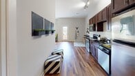 Located along Riverfront Way in Knoxville, Tennessee, this single-floor residence offers a well-designed layout that balances modern style with everyday functionality. The open-concept living area is anchored by rich hardwood flooring that flows throughout the main spaces, complemented by neutral gray walls and a ceiling fan that contribute to a comfortable, airy atmosphere. The kitchen is a standout feature, showcasing deep-toned wood cabinetry, black granite countertops, a classic white subway tile backsplash, and a full suite of stainless steel appliances including a range, microwave, and refrigerator. A built-in wall-mounted bar shelf with seating adds a practical and stylish casual dining option. The living room provides a generous gathering space with high ceilings, track lighting, and a glass-paneled exterior door that introduces natural light and outdoor access. The bedrooms feature plush carpeting, soft neutral tones, and windows dressed with grommet-top curtains and horizontal blinds, offering both comfort and light control. The primary bedroom is notably spacious with vaulted ceiling lines. The bathroom continues the home's clean aesthetic with wood-look flooring and crisp white paneled doors. Overall, the residence presents a cohesive, contemporary design with thoughtful details throughout.