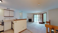 This single-floor residence offers a practical and comfortable layout centered around an open-concept living and kitchen area. The kitchen is well-appointed with white cabinetry accented by warm wood trim, a double-basin stainless steel sink, a dishwasher, a range, and generous counter space. A half-wall peninsula separates the kitchen from the living area, creating a connected yet defined flow between the two spaces. The living room features light carpeting, a tile-surround fireplace, and a large sliding glass door paired with a classic French door that opens to an outdoor deck, filling the space with natural light and providing pleasant outdoor views. The bedrooms are carpeted and bright, each featuring windows with wood trim that frame views of lush greenery outside. The rooms offer a clean, neutral palette with warm wood baseboards and trim throughout. The bathroom includes a vanity with under-sink storage, a wood-framed mirror, and a three-globe light fixture, alongside a stacked washer and dryer conveniently nearby. Consistent warm wood trim details and light-toned walls create a cohesive, welcoming ambiance throughout the entire home.