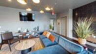 Located at 371 Technology Center Way in Rock Hill, SC, this single-floor amenity space offers a thoughtfully designed environment for relaxation and social interaction. The open-concept layout is anchored by a spacious lounge area furnished with a plush blue sectional sofa, a marble-top coffee table, and a wall-mounted flat-screen TV illuminated by a striking multi-arm gold chandelier. The adjacent entertainment zone features a full-size shuffleboard table set against a dramatic, large-scale abstract art mural that serves as a bold focal point throughout the space. A sleek wet bar equipped with dark cabinetry, a stainless steel under-counter refrigerator, and a wood-paneled accent wall provides convenient beverage service. The dining area accommodates group gatherings with a long wooden table, modern seating, and pendant lighting, all bathed in natural light from expansive windows fitted with horizontal blinds that offer outdoor views. Light hardwood-style flooring flows consistently throughout, complementing the cool gray wall tones. An accessible restroom with dark textured tile accents is conveniently located near the entrance. The overall ambiance is contemporary and inviting, balancing functional amenities with sophisticated, design-forward aesthetics.