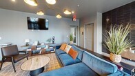 Located at 371 Technology Center Way in Rock Hill, SC, this single-floor amenity space offers a thoughtfully designed environment for relaxation and social interaction. The open-concept layout is anchored by a spacious lounge area furnished with a plush blue sectional sofa, a marble-top coffee table, and a wall-mounted flat-screen TV illuminated by a striking multi-arm gold chandelier. The adjacent entertainment zone features a full-size shuffleboard table set against a dramatic, large-scale abstract art mural that serves as a bold focal point throughout the space. A sleek wet bar equipped with dark cabinetry, a stainless steel under-counter refrigerator, and a wood-paneled accent wall provides convenient beverage service. The dining area accommodates group gatherings with a long wooden table, modern seating, and pendant lighting, all bathed in natural light from expansive windows fitted with horizontal blinds that offer outdoor views. Light hardwood-style flooring flows consistently throughout, complementing the cool gray wall tones. An accessible restroom with dark textured tile accents is conveniently located near the entrance. The overall ambiance is contemporary and inviting, balancing functional amenities with sophisticated, design-forward aesthetics.