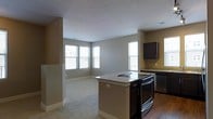 This single-floor space showcases an open-concept layout that seamlessly connects the kitchen and living area. The kitchen is well-appointed with dark cabinetry, granite countertops, and a suite of black appliances, including a range and a stainless steel dishwasher. A central island with a matching granite surface provides additional workspace and serves as a natural dividing point between the kitchen and the adjoining living area. The kitchen features hardwood flooring, which transitions to plush carpeting in the living area, creating a clear yet cohesive distinction between the two spaces. Neutral-toned walls in a warm beige complement the overall design, lending a clean and understated aesthetic throughout. Multiple windows along the exterior walls allow generous natural light to fill the space, enhancing its open and airy feel. The living area offers a comfortable, versatile footprint suitable for a variety of furniture arrangements. The overall design balances functional kitchen amenities with a welcoming living environment, making the space practical and visually cohesive.