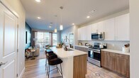 Located at Bravery Heights in Colorado Springs, CO, this thoughtfully designed 2-bedroom, 2-bathroom residence offers a cohesive blend of modern style and functional living. The open-concept kitchen and living area forms the heart of the home, anchored by a large center island with a white quartz countertop, bar seating, and a striking two-tone cabinet design pairing crisp white upper cabinets with warm wood-toned lower cabinets. Stainless steel appliances, including a gas range, microwave, refrigerator, and dishwasher, complete the well-appointed kitchen. Light wood-style flooring flows throughout the main living areas, complementing the neutral color palette. The living room features a bold teal accent wall, large sliding glass doors that invite natural light, and access to an outdoor balcony. The primary bedroom is a comfortable retreat with plush carpeting and direct access to its en-suite bathroom, which includes a dual-sink vanity with wood cabinetry, a glass-enclosed shower, and a walk-in closet. The second bedroom is well-sized with bright windows and a ceiling fan. Both bedrooms offer dedicated closet space. The overall ambiance is contemporary and warm, with consistent finishes tying every room together seamlessly.