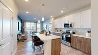 Located at Bravery Heights in Colorado Springs, CO, this thoughtfully designed 2-bedroom, 2-bathroom residence offers a cohesive blend of modern style and functional living. The open-concept kitchen and living area forms the heart of the home, anchored by a large center island with a white quartz countertop, bar seating, and a striking two-tone cabinet design pairing crisp white upper cabinets with warm wood-toned lower cabinets. Stainless steel appliances, including a gas range, microwave, refrigerator, and dishwasher, complete the well-appointed kitchen. Light wood-style flooring flows throughout the main living areas, complementing the neutral color palette. The living room features a bold teal accent wall, large sliding glass doors that invite natural light, and access to an outdoor balcony. The primary bedroom is a comfortable retreat with plush carpeting and direct access to its en-suite bathroom, which includes a dual-sink vanity with wood cabinetry, a glass-enclosed shower, and a walk-in closet. The second bedroom is well-sized with bright windows and a ceiling fan. Both bedrooms offer dedicated closet space. The overall ambiance is contemporary and warm, with consistent finishes tying every room together seamlessly.